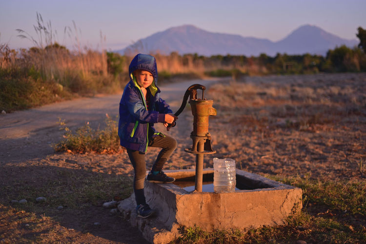 Boy standing by water pump on field against sky