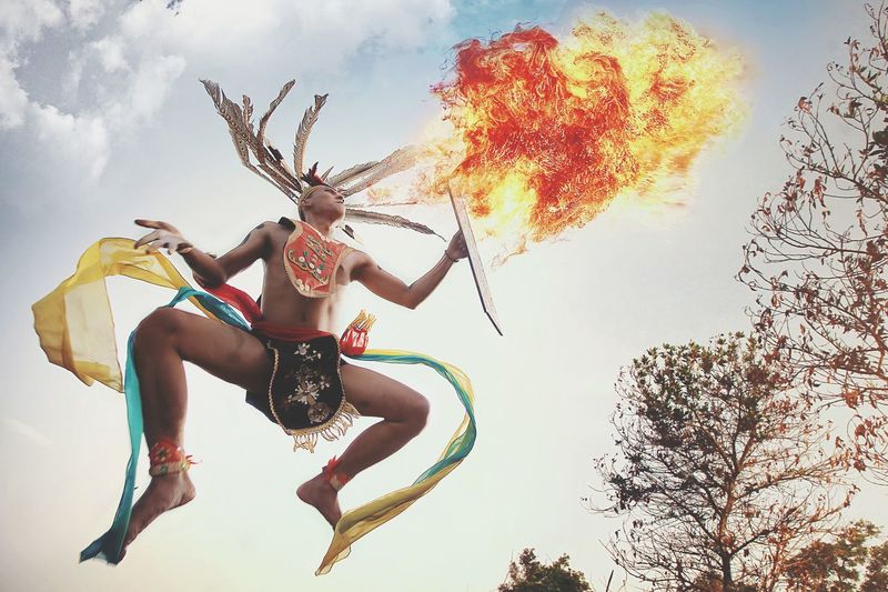 Low angle view of man wearing traditional clothing blowing fire against sky