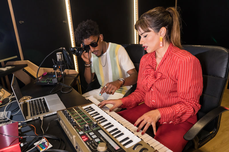 Female musician playing electric piano and black male singer listening to melody in headphones while recording music in studio