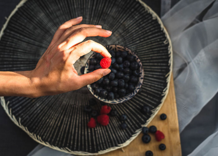 High view of a woman's hand holding a raspberry. underneath is a little bowl more blueberries.