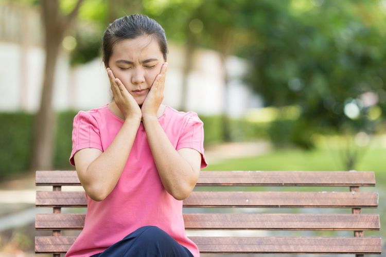 Young woman suffering from toothache sitting on bench at park