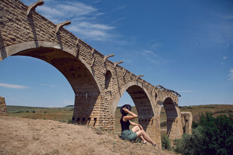 Woman traveler explorer in a black hat sit next to the destroyed old stone bridge in the summer