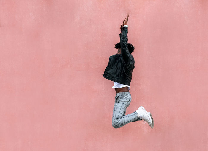Mid adult man with arms raised jumping by pink wall in city