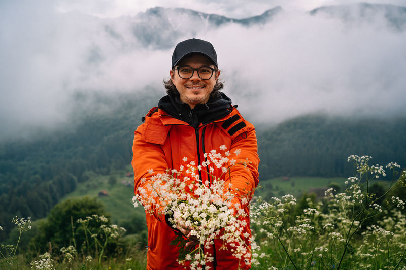 Smiling man standing on field and holding wildflowers  against misty mountain in summer