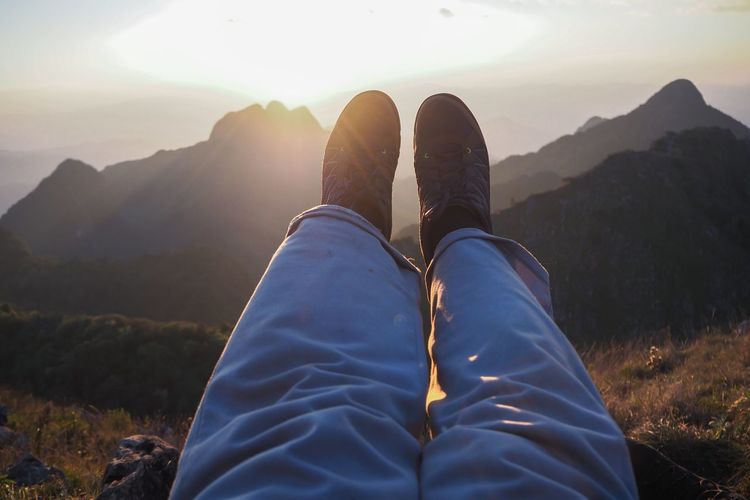 Low section of person relaxing on land against mountains during sunset