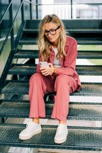 Young casual woman using smartphone while sitting on the iron steps of a modern office building.