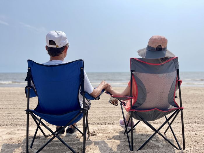 Senior couple affectionally holding hands  seated on beach against water and sky.  rear view.    
