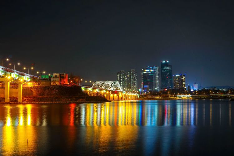 Illuminated buildings by river against sky at night