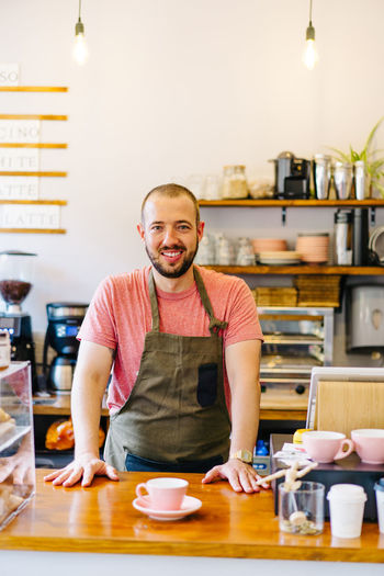 Cheerful male barista in apron smiling and looking at camera while standing at counter with cup of coffee in coffeehouse