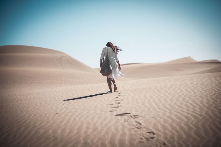 Rear view of woman walking on sand dune at desert against clear sky during sunny day