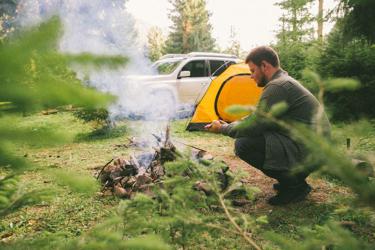 Man build fire in forest, car with yellow tent on background. camping