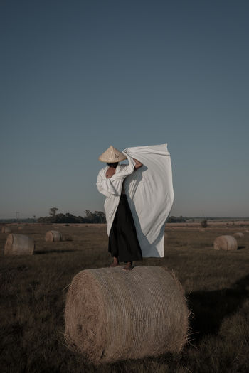 Rear view full length of young man holding fabric while standing on hay bale at agricultural landscape