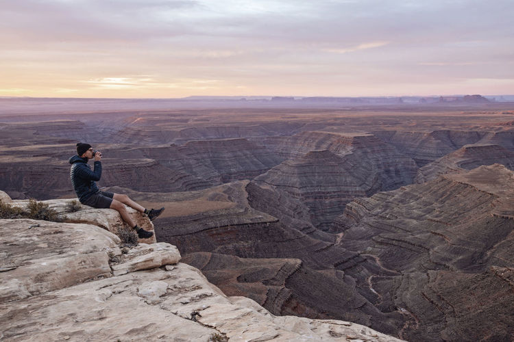 Man drinks coffee sitting on edge of cliff, mexican hat, utah