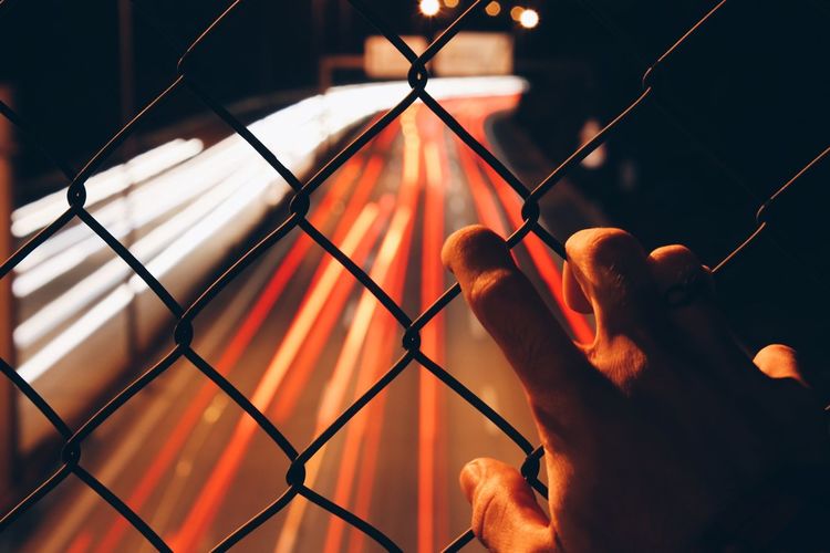 Cropped hand holding chainlink fence against light trails at night