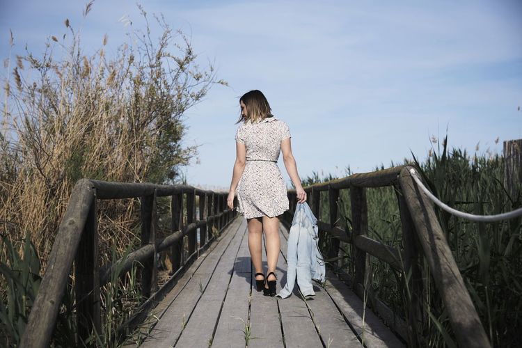 Full body back view of young female in casual dress with jacket in hand strolling on wooden footbridge among tall dry grass in sunny day