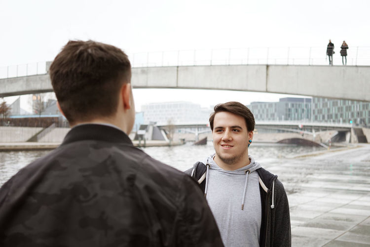 Man looking at friend by river with bridge in background