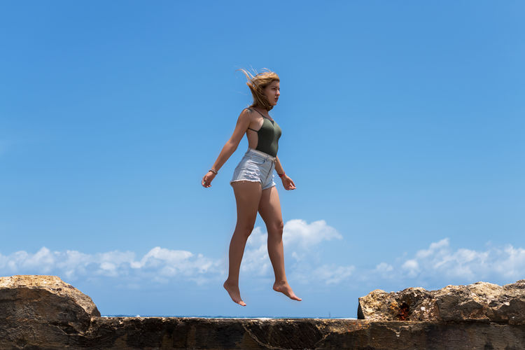 Low angle view of woman jumping on seawall against blue sky