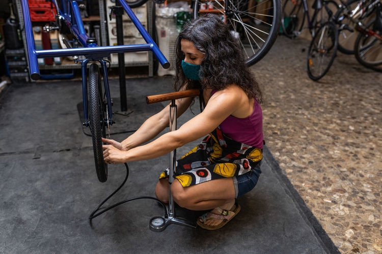 Full length of adult ethnic female mechanic inflating bicycle tire with pump while working in professional workshop
