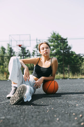 Portrait of a charming girl sitting on a sports field in a park or school with a basketball