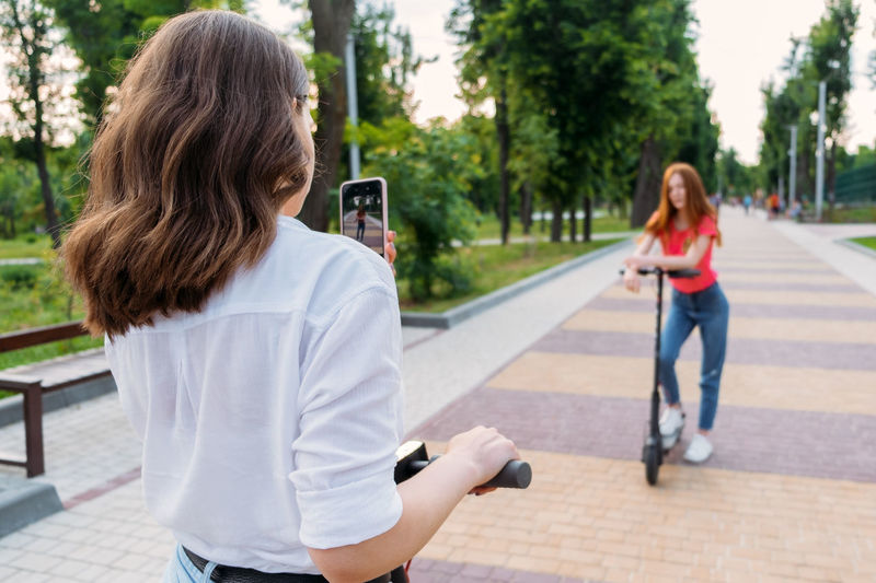 Two girl friends riding push e scooter and do selfie via their smartphones outdoors. group gen z