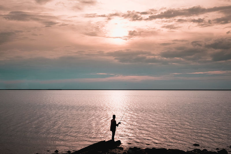 Silhouette man fishing in sea against cloudy sky during sunset