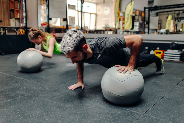 Full length of strong determined athletic man and woman doing plank exercise using one hand with medicine ball during workout in gym