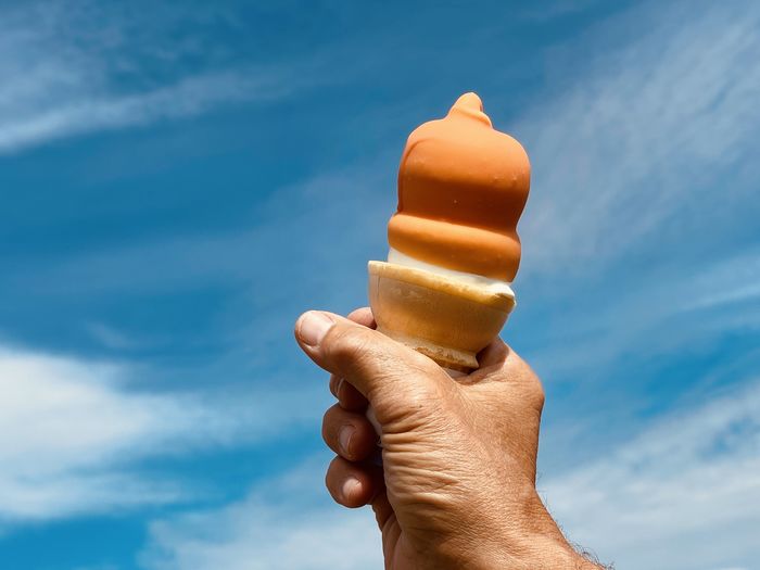Cropped mans hand holding orange ice cream cone against blue sky.