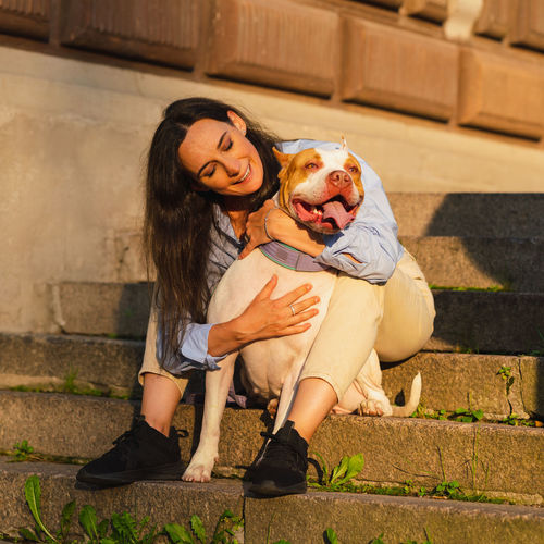 Woman sitting on stone staircase and cuddling with happy american pitbull terrier