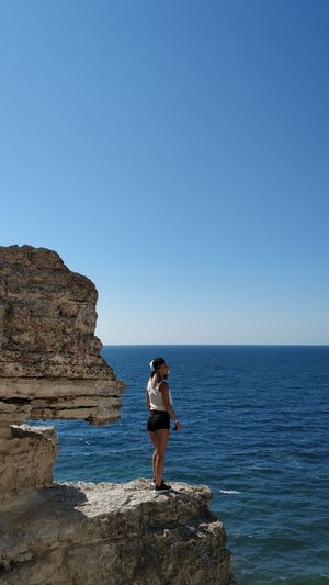 Woman standing on rock formation by sea against clear blue sky