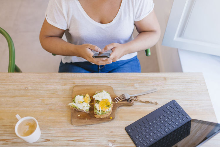 Top view of a girl having breakfast and typing with a smartphone. she is working from home.