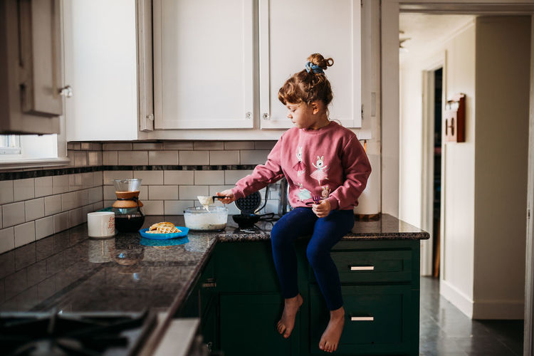 Young girl sitting on kitchen counter scooping waffle batter