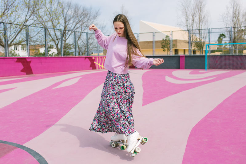 Charming young female in colorful skirt and lilac sweatshirt standing on quad roller skates on pink playground in spring day in city