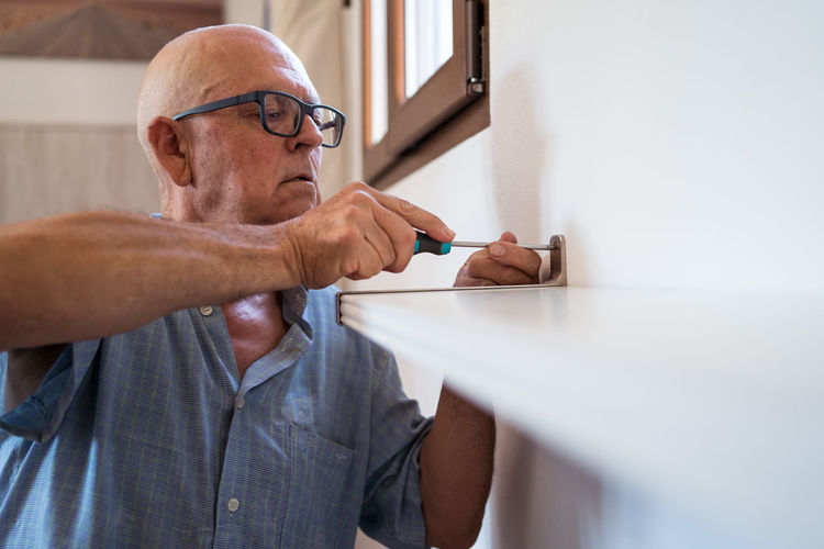 Attentive elderly male in eyeglasses with manual screwdriver screwing shelf to wall in house room