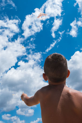 Rear view of shirtless boy flying kites against sky