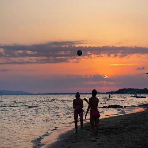 Friends playing volleyball at beach against sky during sunset