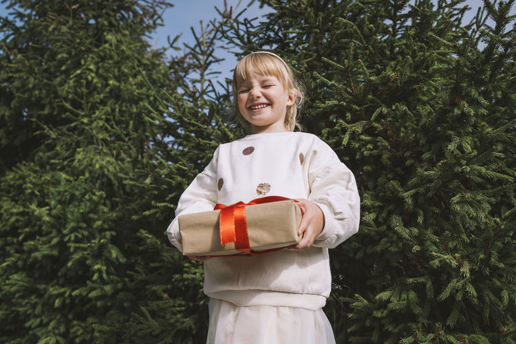 Smiling girl with gift box standing in front of fir tree