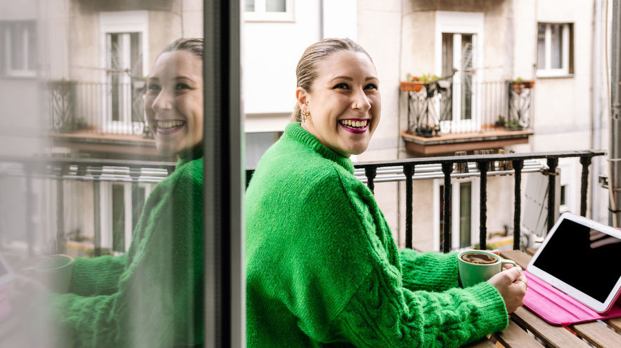 Smiling woman is drinking coffee on her balcony while using a tablet.
