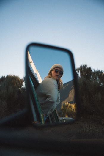 Reflection of young woman on side-view mirror against clear blue sky