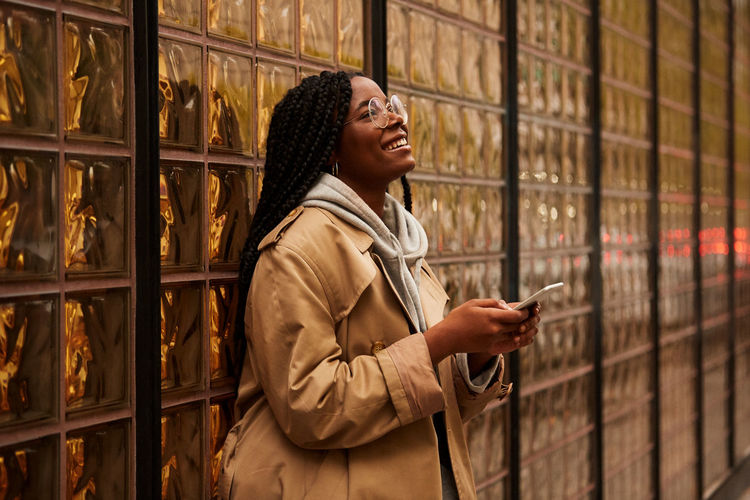 Confident black female with braids in casual outfit looking away and browsing smartphone while standing near glass wall