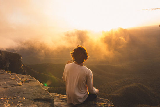 Rear view of man sitting on cliff while looking at landscape during sunset