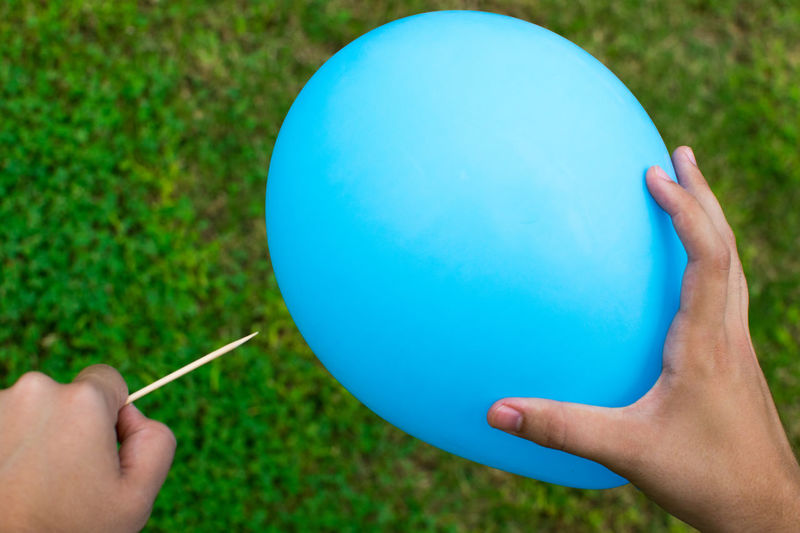 Cropped hands holding toothpick and blue balloon over lawn