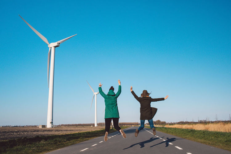 Full length of two girls jumping on road against a wind turbine  and clear sky