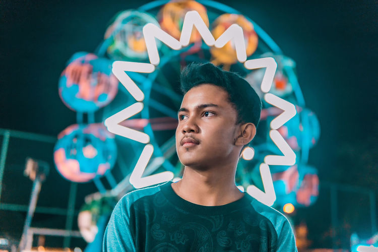 Young man looking away against illuminated ferris wheel at night