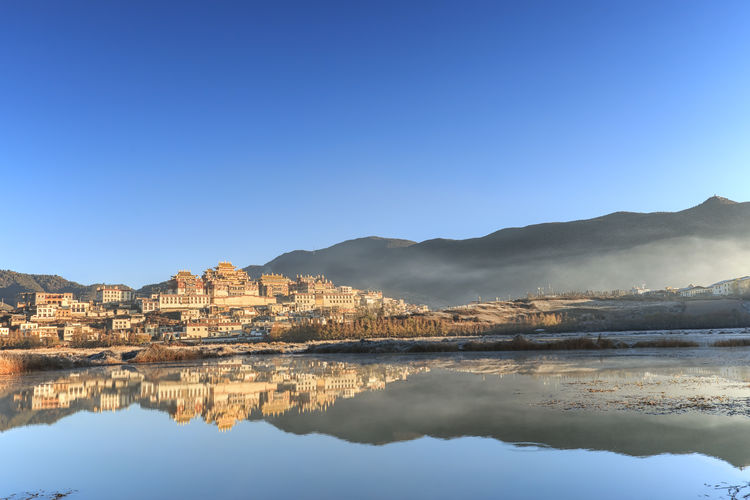 Reflection of ganden sumtseling monastery on lake against clear blue sky