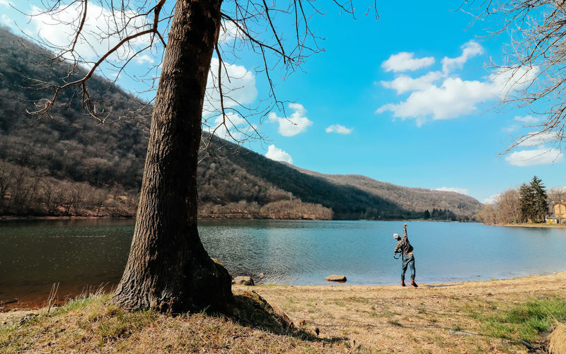 Man standing on tree by lake against sky