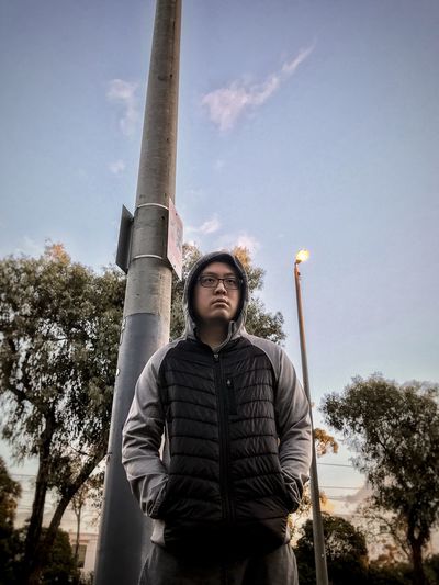 Low angle view of man in hoodie looking at camera against lamp post and sky.