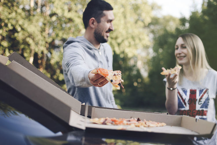 Couple eating pizza while standing against trees at park