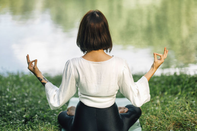 Young woman sitting in lotus position and practicing meditation near water in the nature