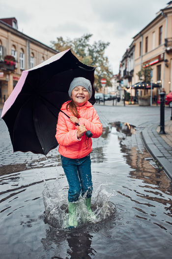 Happy smiling girl holding big umbrella jumping in the puddle during walk in a downtown on rainy day