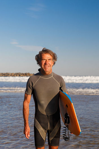 Smiling man holding surfboard standing on beach against sky